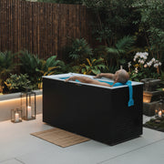 Person lying in a modern outdoor cold plunge tub with plants and a wooden deck in the background